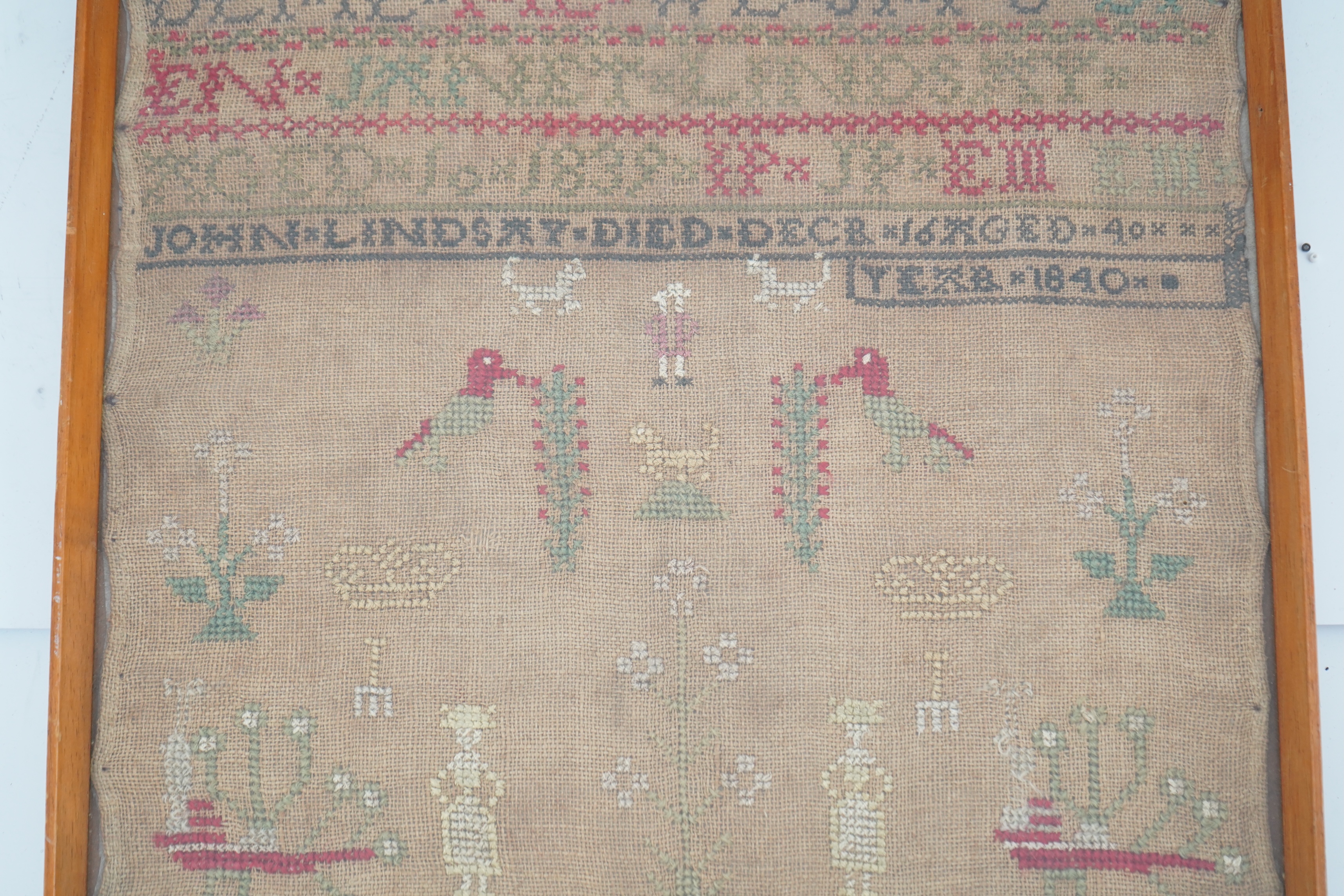 An alphabet cross stitch sampler with a figurative lower spot motif panel and commemorative dedication to John Lindsay, died Dec 16 1840, 27cm wide x 43.5 cm high. Condition - some light staining to linen, the blue and g